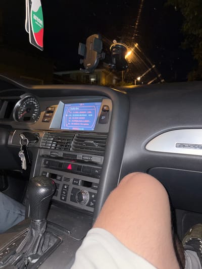 The image shows the interior of a car, specifically the dashboard area. The steering wheel and leg of a person are visible. The car's console displays radio information, which reads in Bulgarian. The gear shift is visible. A phone mount and air freshener hang on the window. A cluster of keys is in the ignition. Text indicates that the car is an Audi. The time displayed on the screen is 22:46.
