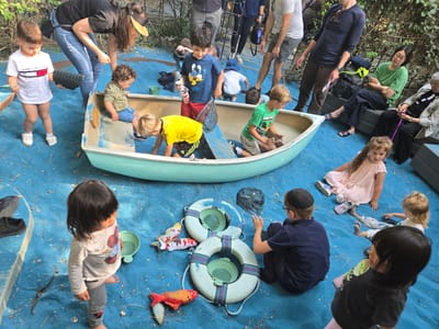 A group of children and adults are gathered around a light blue canoe on a blue surface, possibly a painted playground surface. The canoe is centrally located. Several children are inside the canoe, while others are around it, both on the blue surface and on the surrounding green grass or foliage. Some adults are standing near the canoe and interacting with the children. There are various toys and props scattered around, including life preservers and stuffed fish. The scene appears to be outdoors, possibly in a park or schoolyard. Based on the presence of foliage and architectural elements, the location may be in Pasadena, United States of America.