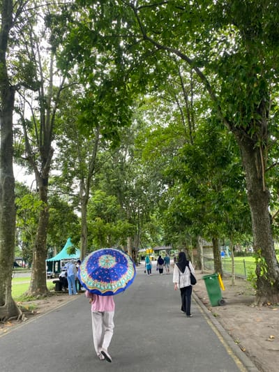 The scene is a paved walkway lined by lush green trees. It is a partly cloudy day. Seven people are walking along the walkway, most facing away from the camera. A person with a blue and white umbrella is walking towards the camera, obscuring the person's face. One person is wearing a black hijab and walking in the same direction as the person with the umbrella. A yellow trash can is visible on the right side of the pathway. There is a small, teal tent-like structure on the left, and a grass patch is visible to the left of the walkway.