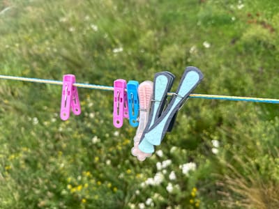 A clothesline is strung horizontally across the frame, with several colorful clothespins attached. The clothespins are a mix of bright pink, blue, and pale pink and light blue, with the pink and blue ones having a more traditional design and the pale ones appearing to be a softer, more rounded shape. Some of the clothespins, particularly the lighter colored ones, appear to have water droplets on them. The clothesline itself is a braided rope of yellow and blue.

The background is a soft, blurred expanse of green foliage, dotted with small white and yellow wildflowers. This suggests an outdoor setting, likely a garden or a grassy field. The bokeh effect of the background indicates a shallow depth of field, with the focus strongly on the clothespins and clothesline.

The scene is bathed in natural light, suggesting daytime, but the overall mood is calm and perhaps slightly damp due to the water droplets. There is no visible text in the image. The composition is simple, focusing on the everyday objects of clothespins against a natural backdrop. The location context of Tuishevo, Russia, suggests this scene is taking place in a rural or semi-rural environment.