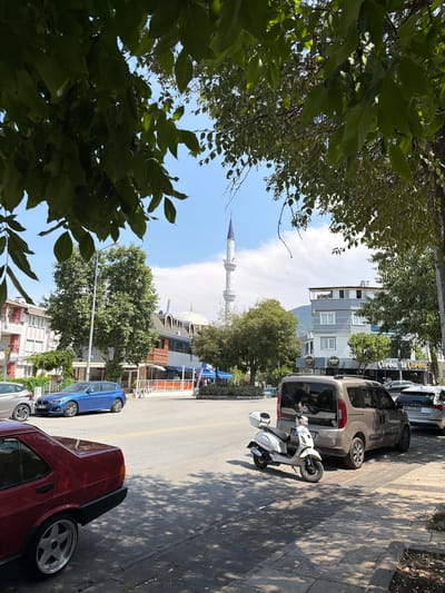 In Denizli, Turkey, on a sunny day, a street scene unfolds under a clear blue sky with scattered clouds. The foreground is dominated by the rear of a dark red sedan, its metallic paint reflecting the sunlight and casting shadows. To its right, a white scooter is parked on the sidewalk. Behind the scooter, a beige van is parked on the street. Further down the street, a bright blue hatchback is visible. Buildings line the street, with a mosque's minaret prominently visible in the distance, alongside buildings featuring arched windows and domes. Trees with lush green foliage cast dappled shadows on the street and sidewalk. A sign for "Corba a Corba" is visible on one of the buildings, indicating a food establishment. The road has yellow lane markings.