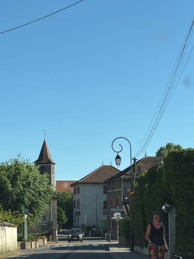 A street scene in the city of Villages du Lac de Paladru, France, shows a vehicle driving down a paved road lined with buildings and lush green foliage under a clear blue sky. A church spire with a weather vane is visible to the left, while buildings with red-tiled roofs and a street lamp adorn the right side of the road. A woman in a black top and colorful patterned pants walks on the right side of the road. Signs with French text are posted along the street.