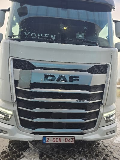 A white DAF 480 truck is depicted in a frontal view, stationary on a paved surface. The vehicle's prominent grille features horizontal chrome and black slats, with the "DAF" logo centered and "480" visible on the upper right side. Its modern LED headlights are illuminated. A Belgian license plate, "2-GCK-043," is affixed to the lower front bumper. The windshield reflects the bright sky and shows a white banner inside with the text "YOHEN" and a small hanging ornament near the center. The ground beneath the truck is a dark, patterned surface, possibly pavement, partially covered with melting snow or slush, exhibiting tire tracks. No individuals are visible, nor are any actions or interactions occurring. The environment appears to be an outdoor, open area. The location is Ostend, Belgium.