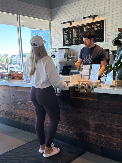 A blonde woman wearing a white baseball cap, a white sweatshirt, and dark purple leggings stands facing a dark wood counter in a coffee shop. She is looking at the menu board above the counter. A man with dark curly hair, wearing a dark t-shirt, stands behind the counter, appearing to take her order. Bags of potato chips are stacked on the counter in front of him. The coffee shop has a tiled white wall and a large window overlooking a city street. The scene appears to be in Edgewater, United States of America.