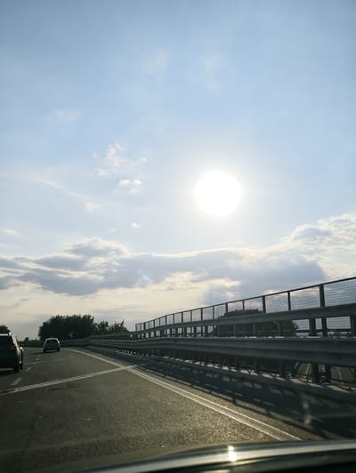 The image captures a view from a moving vehicle on a highway, likely in or near Marigliano, Italy, given the context. The sun is bright and high in the sky, with scattered clouds. A metal guardrail with a mesh fence runs along the right side of the road, indicating an elevated position, possibly a bridge or overpass. Trees are visible in the distance beyond the guardrail. Two cars are on the highway, with one in the foreground slightly to the left and another further ahead in the center lane. The road surface is asphalt with white lane markings. The overall scene suggests a daytime journey on a well-maintained road.