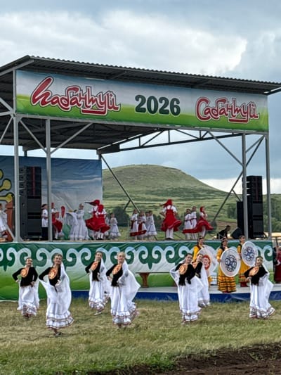 The image captures an outdoor performance on a stage in the village of Nurkeevo, Russia, likely during an event or festival. A large banner with stylized text and the year "2026" is prominently displayed above the stage. On the stage, a group of young women in traditional white and red dresses are performing a dance. Some are holding round, decorated shields. In the background, a hilly, green landscape under a cloudy sky is visible. In the foreground, another group of young women, also in traditional attire, some with white veils and black tops with gold accents, are positioned lower than the stage, suggesting they are part of the event or audience. The overall atmosphere appears festive and cultural.