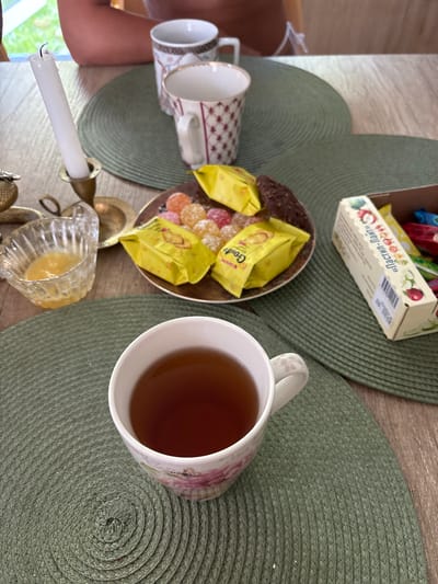 In Vardane-Verino, Russia, a table is set with a tea service. Two decorative cups sit on a green woven placemat. One cup is empty, the other is filled with dark tea. To the right of the cups is a plate of candies and individually wrapped sweets. To the left of the cups, a white candle in a brass candlestick stands upright. A small glass bowl containing a yellow substance, possibly honey, is also on the table. The table is made of light-colored wood and a person's arm is visible in the background, suggesting a relaxed, domestic setting.