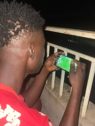 An overhead shot captures a person, likely male, holding a smartphone in both hands and looking intently at its screen. The individual is positioned outdoors, against a very dark background that suggests nighttime, possibly on a balcony or elevated structure, indicated by a light-colored, rusty metal railing in the foreground and stretching across the mid-ground.
The person has short, dark, textured hair (appearing as dreadlocks or twists) and is wearing a red short-sleeved shirt with some indistinct white graphics visible on the right side. Their skin tone is dark.
The smartphone screen is brightly illuminated, displaying what appears to be a mobile football (soccer) game. The screen clearly shows a green pitch with white markings, including a penalty box, center circle, and corner arcs, along with small white and blue player icons or indicators. The person's thumbs are positioned as if actively playing the game.
Notable details include the heavy shadows and dark environment, indicating low ambient light conditions. The person's head is tilted upwards, focusing entirely on the phone. No clear text is legible on the phone screen or clothing. The scene takes place in Abatete, Nigeria.