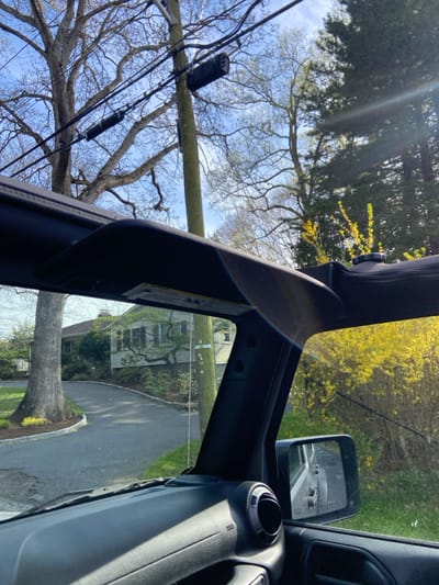 The image was taken from inside a black Jeep Wrangler, looking out the driver's side window, with the sun shining brightly through the front windshield. The driver is parked on a paved driveway, facing the street. There is a large tree on the driver's side of the driveway, with green grass and a lush lawn on the other side. A one-story house is visible on the right side of the driveway. The location is Rye, New York, United States (Latitude 40.954, Longitude -73.709). The image shows a rear-view mirror, a dashboard with a vent, a sunshade, and the top of a vehicle's door. The sun's reflection on the windshield and windows are clearly visible. The image is well-lit, with a bright blue sky and sunshine.