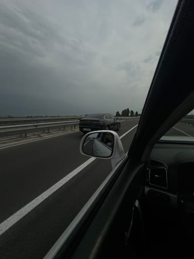 The image is a first-person perspective shot from inside a car, looking out through the passenger side window and side mirror. The car is on a highway with multiple lanes marked by white lines. The road surface is asphalt. A dark-colored car is visible ahead, also on the highway. The side mirror reflects the interior of the car, showing a person's hand with a ring, and the exterior, showing the road and another car. The sky is overcast and cloudy, suggesting a potentially gloomy or rainy day. There are trees lining the side of the highway in the distance, and a concrete barrier separating the lanes. The interior of the car shows a black dashboard with air vents. The image is oriented vertically, making the highway appear to run up the frame. No specific time of day can be determined, but the lighting suggests daylight. The location is Sergeli, Uzbekistan. There is some text on the license plate of the car ahead, but it is too blurry to read.
