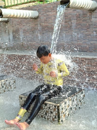 A young boy is sitting on a stone bench, getting splashed with water from a pipe overhead. The pipe is attached to a wall made of brick and stone, and there is a second pipe running horizontally above it. The boy is wearing black pants, a yellow shirt with a white pattern, and a blue wristband. He is smiling as he gets wet. The water is spraying all around him, creating a misty effect. The scene is set in a water park in Kecamatan Tulung, Indonesia. The coordinates for this location are latitude: -7.592, longitude: 110.65.