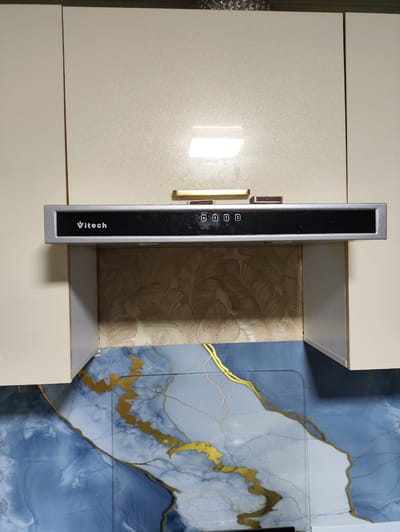 The image shows a close-up, eye-level view of a kitchen range hood mounted between two cream-colored cabinets. The range hood has a silver-toned metallic frame and a black control panel with white lettering that reads "Vitech". Below the control panel, there are several illuminated buttons labeled with symbols and numbers 1, 2, and 3. Two unlit matchsticks are placed on the top edge of the range hood, one to the left of the center and another to the right. The backsplash behind the range hood features a textured wallpaper with a pattern of stylized leaves in beige and gold tones. The lower part of the image shows a countertop or backsplash with a blue and white marbled pattern accented with gold veining. The scene is indoors, likely in a kitchen. The location is identified as Navruz, Uzbekistan, but no specific landmarks or environmental details in the image support this location.