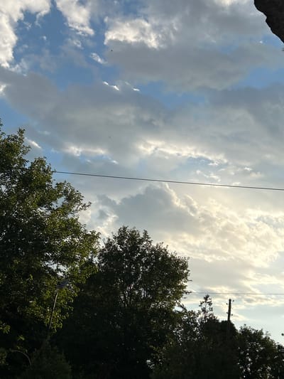 The image captures a segment of the sky with scattered clouds, illuminated by sunlight. In the foreground, the silhouettes of trees are visible, with their dark green foliage contrasting against the bright sky. A thin wire runs horizontally across the frame, obscuring some of the cloud formations. On the right side of the image, a telephone pole with insulators is partially visible above the treeline, extending towards the sky. The overall scene depicts an outdoor environment, likely a suburban or rural area, at an unspecified time of day, judging by the quality of light. The location of Marino pole, Bulgaria is not explicitly represented in the visual elements of the image.