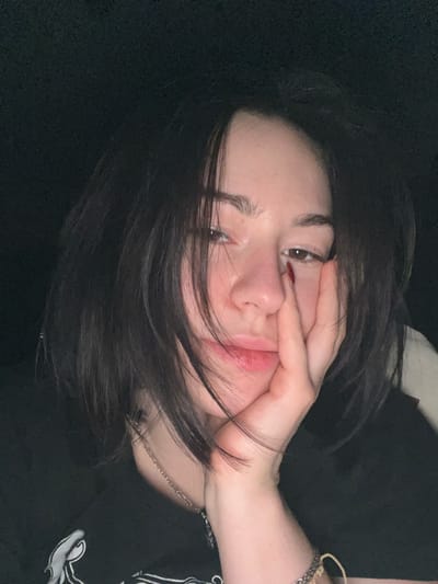 A person with dark hair and fair skin poses close to the camera with their hand on their face. They have brown eyes, full lips, and appear to be wearing a black t-shirt. A silver necklace is visible around their neck, and bracelets adorn their wrist. The background is mostly black, adding to the overall dark and intimate feel of the portrait.