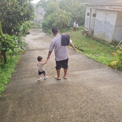 An adult male, wearing a light-colored shirt, dark pants, and a black bag over his shoulder, is walking on a concrete pathway, holding the hand of a small child. The child is wearing a light-colored shirt and dark pants. The scene is located in Kecamatan Tamansari, Indonesia, at coordinates -6.635 latitude and 106.737 longitude. The pathway is lined with lush green foliage and there are several buildings visible in the background. The buildings appear to be residential homes with a plain exterior.  The man is walking in the direction of the buildings. The child is looking at something off-camera to the left, while the man is focused on the path ahead. There is a general sense of calm and normalcy in the scene.