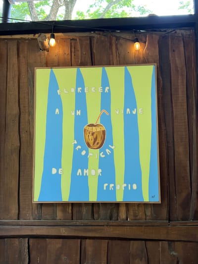 The image shows a framed painting mounted on a wooden wall. The painting depicts a coconut with a straw inserted into it, surrounded by blue and green vertical stripes. The text "FLORECER A UN VIAJE TROPICAL DE AMOR TROPIO" is written in white letters around the coconut. The painting is signed "AIP" in the bottom right corner. The wall is made of brown wood planks and there are two light bulbs with wires attached, one on either side of the painting. The location of the image is based on the coordinates provided, which are 9.643 latitude and -85.167 longitude, and correspond to the city of Santa Fe, Costa Rica. There are no people or other objects in the image.