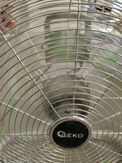 The image shows a close-up, head-on view of the front grill of an electric fan. The grill is made of silver metal, with concentric circular wires and radial wires. The fan blades and motor housing are visible behind the grill, blurred due to motion or shallow depth of field. A black circular emblem with a white gear and the word "GEKO" is present at the center of the grill. The background is a light, neutral color, with some indistinct shapes and colors suggesting an indoor environment. There are no people or animals visible. The scene is static, with no discernible action. The image is a still photograph. The provided location of Palmiry, Poland, is not visually verifiable from the image content.