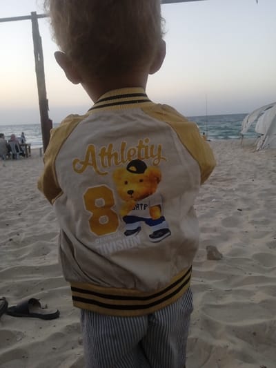 A young child, viewed from behind, stands on a sandy beach in Al-Mawasi, Palestinian Territories, looking towards the sea under a twilight sky. The child has light-colored, curly hair and wears a light-beige bomber jacket with yellow sleeves and black-striped cuffs and hem. The jacket's back features "Athletiy" in yellow script, a large yellow number "8," and an illustration of a brown bear wearing a baseball cap, a white t-shirt with "SHTP," blue pants, and black and white shoes. The child also wears light blue and white striped pants. To the child's left, a dark-colored sandal rests on the sand. In the mid-ground to the left, several individuals are seated around a table on the beach, near a vertical wooden pole. Further right, a person stands by the water's edge, engaged in fishing. White tent-like structures are partially visible on the sand to the right. The sea exhibits gentle waves under a pale, gradient sky.