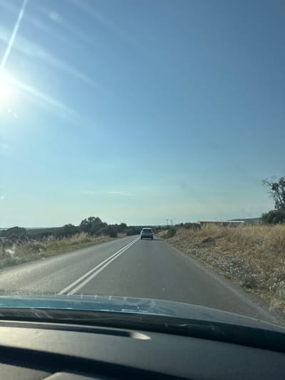 The image shows a view from the perspective of a driver on a two-lane asphalt road. The road has white double lines down the center. In the distance, a silver car is driving away from the viewer. The landscape on both sides of the road consists of dry, brown grass and scattered bushes and trees. The sky is clear blue with bright sunlight creating a lens flare effect in the upper left corner. The foreground shows the dashboard and windshield of the car from which the photo was taken. The location is identified as Nea Triglia, Greece.