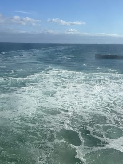 This image captures a choppy sea with white foam churning in the foreground. The water transitions to a darker blue in the distance, meeting a hazy horizon under a bright blue sky with scattered white clouds. In the middle distance, on the right side of the frame, a dark, rectangular structure, possibly a pier or breakwater, juts out into the sea. The scene suggests a possibly windy day at sea, with no people or specific landmarks clearly visible to identify a precise location within Unknown, Denmark.