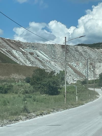 In Kazreti, Georgia, a wide, paved road curves gently to the right, its surface appearing dusty or unpaved in some areas. To the left of the road, a steep, barren hillside dominates the landscape. This hillside is composed of various types of rock and soil, exhibiting distinct striations of light and dark colors, suggesting a mined or excavated area. Scattered trees and vegetation grow at the base of the hillside and in the foreground. Several utility poles with wires are visible along the roadside and extending towards the hillside, with one pole featuring a lamppost. The sky above is blue with scattered white cumulus clouds.