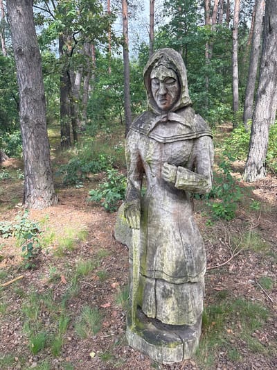 A weathered wooden sculpture of a woman stands in a forest clearing. The sculpture depicts a figure wearing a hooded garment, with carved facial features and textured clothing. The woman appears to be holding a staff or pole in her left hand and has her right hand clasped in front of her. The sculpture is set amidst trees with brown bark and green foliage, with a ground cover of dry leaves, pine needles, and grass. The background consists of more trees and undergrowth, suggesting a natural, outdoor environment. The image was taken in Sielachowskie, Poland.