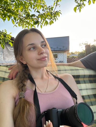 A young woman with light brown hair braided down her back and wearing a pink ribbed tank top is taking a selfie outdoors. She is sitting on a checkered couch next to a person whose arm is visible resting on her shoulder. The woman is holding a black Sony camera with a green lens filter. Behind them, a house with a dark tiled roof is visible, along with a chain-link fence and trees. The scene is lit by the warm glow of the setting sun, casting long shadows. The location is identified as Yablunivka, Ukraine.