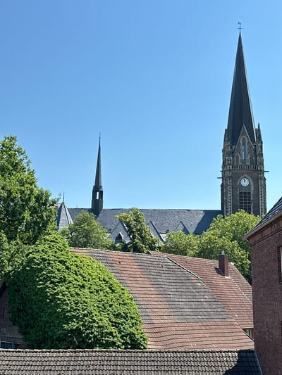 This media file depicts an outdoor scene featuring a church spire and adjacent buildings. The church spire, constructed from stone, is tall and pointed, topped with a cross. A clock face is visible on the side of the church tower. The surrounding structures include tiled roofs and brick facades. Lush green trees with dense foliage are interspersed throughout the scene, partially obscuring some of the buildings. The sky is a clear, bright blue, suggesting a sunny day. The perspective is from an elevated viewpoint, looking down and across the scene. There are no people visible. The overall impression is one of a historic urban or town setting. The time of day appears to be daytime, likely morning or afternoon, due to the clear sunlight and shadows cast. The location is specified as Recklinghausen, Germany. No text is visible in the image.
