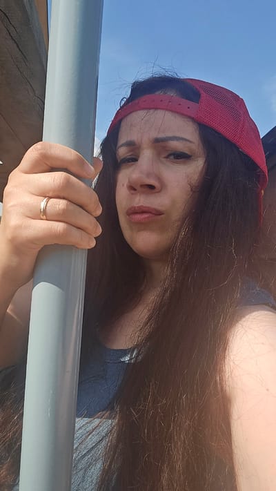 This media file shows a close-up, rotated selfie of a woman outdoors. She has long, dark hair and is wearing a red baseball cap. Her left hand, adorned with a ring, is resting on a grey bar, which appears to be part of a railing or structure. Her facial expression is neutral, with her lips slightly parted and her eyes looking towards the camera. The background suggests a bright, sunny day, with a clear blue sky visible. The overall lighting indicates it is daytime. The scene is set outdoors, with the location context provided as Ермолаевский Затон, Russia. There is no visible text in the image.