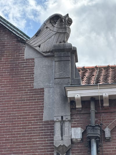 The image shows a close-up of the facade of a building in Roermond, Netherlands. The building is constructed of red brick with stone architectural elements. At the top left, a green tiled roof is visible. Dominating the upper part of the image is a stone carving of an owl with its wings spread, perched on a stone pedestal. Below the owl, a large, grey stone architectural feature is attached to the brick wall. The image also shows a section of a tiled roof and a drainpipe. The sky is cloudy.