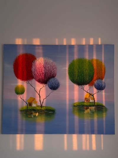 A rectangular painting hangs on a light gray wall. The painting depicts two scenes, one above the other, like a reflection. Each scene features a grouping of round, fluffy trees in vibrant colors (orange, green, red, purple, blue) connected by branches to a small, stylized house on a hill. The scenes are mirrored, with the upper scene inverted relative to the lower. The background is a soft blue with subtle streaks of pink, suggesting a sky. Light and shadow from a window are visible on the wall to the right of the painting, creating horizontal bars of light. There is no indication that the scene is in Ariel, Palestinian Territories.