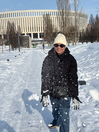 A female subject stands centrally in the foreground of a snow-covered outdoor scene, smiling directly at the viewer. She wears a white knit beanie, black sunglasses, a black puffer jacket, blue jeans, and white/grey gloves. A black fanny pack is visible across her torso. Snow particles are suspended in the air around her, adhering to her jacket and gloves, with a visible accumulation on her left glove and scattered particles around her, consistent with snow being thrown or disturbed. The ground is covered in deep, white snow, with a cleared, snow-dusted path leading into the background. Numerous bare deciduous trees are interspersed throughout the park-like environment. In the background, a large, modern, light-colored circular or oval stadium structure with vertical window slits is prominent, identifiable as the Krasnodar Stadium. A small green utility vehicle is discernible on the path further in the background. The sky is clear and blue, and strong sunlight casts distinct shadows on the snow. The scene is located in Plodorodny, Russia.