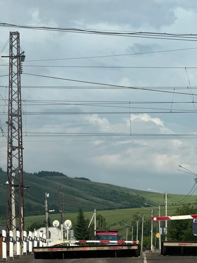 The image depicts a railway crossing in the city of Zykovo, Russia. The foreground shows a raised railway barrier, painted with red and white stripes, blocking a road. A red truck is visible beyond the barrier, waiting to cross the tracks. Overhead, a complex network of electrical wires crisscrosses the sky, supported by tall metal pylons and wooden poles. In the background, rolling green hills under a cloudy sky form the natural landscape. The overall scene suggests a typical day at a rural or semi-rural railway crossing.
