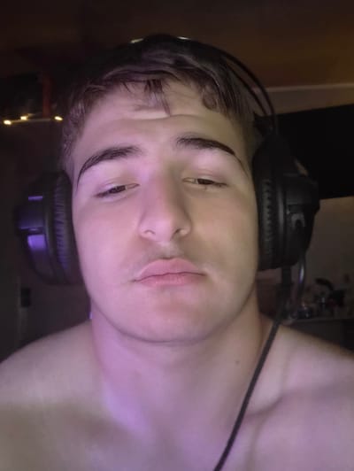 A close-up, head-and-shoulders, low-angle shot shows a young Caucasian male wearing black headphones. He has dark, messy hair and a neutral expression with his lips slightly parted. The lighting is dim, with a purple hue cast on his face and shoulders. The background is dark and out of focus, with some indistinct shapes that could be furniture or objects on shelves. There are no visible signs of the city of Aleksandrovo, Bulgaria in the image.