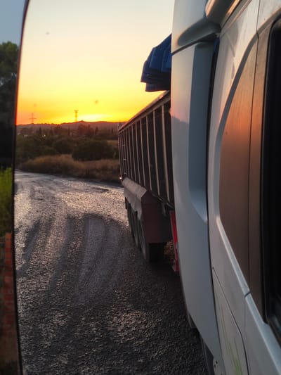 The image captures a sunset view from the perspective of a vehicle, possibly a truck, driving on a rural dirt road in Riba-roja de Túria, Spain. The road is wet and reflects the warm colors of the sky. To the right, the side of a white truck cabin is visible, with its metallic trailer extending into the distance. In the background, a hazy landscape features hills, vegetation, and a power pylon against the vibrant orange and yellow hues of the setting sun. The reflection in a mirror on the left side of the frame shows more of the surrounding environment.