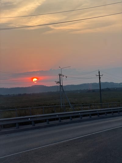 The image captures a sunset over a rural landscape, possibly in Конец-Бор, Russia, as suggested. The sun, a vibrant orange-red orb, is partially obscured by a cloud bank and is positioned low on the horizon, casting a warm glow across the sky. The sky itself transitions from a soft pink and orange near the sun to a pale yellow and then a muted greyish hue at the top. Two thin, dark power lines stretch horizontally across the upper portion of the frame. Below the sky, a silhouetted line of trees marks the distant treeline of a hill or mountain range. In the middle ground, a field of dry grass or crops extends towards the horizon, bordered by a metal guardrail. Several utility poles, one with an attached lamp post, are visible in the middle ground, connected by a network of wires. The foreground consists of a paved road, angled diagonally from the bottom right to the upper left of the image, with a white lane marker. The overall scene conveys a sense of tranquility and the end of the day in a semi-rural setting.