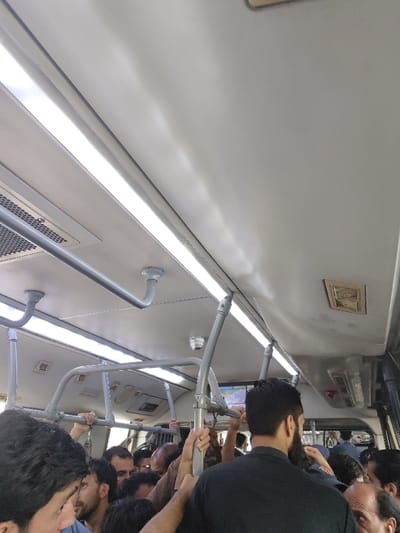 This image depicts the crowded interior of a Metro Bus in Rawalpindi, Pakistan, viewed from a passenger's perspective looking towards the front or middle of the vehicle. The bus is occupied by numerous adult males standing shoulder-to-shoulder, indicating significant passenger density.

The ceiling is light grey and features several long, illuminated LED or fluorescent light fixtures running lengthwise. Below these lights, a system of grey metal handrails and grab bars is installed for standing passengers, many of whom are utilizing them. Visible ceiling components include a rectangular air vent grille on the left and another square vent and a small, beige, rectangular air vent cover on the right. A white, dome-shaped CCTV security camera is mounted on the ceiling structure. Towards the rear of the visible area, a small, illuminated digital display screen is partially visible above passengers' heads.

The passengers are primarily adult males of varying ages, dressed in casual clothing. Many individuals have dark hair and facial hair. Their faces are partially obscured by the density of the crowd and the camera's angle, but several individuals are observed looking forward. No distinct individual interactions are discernible beyond the collective presence within the confined space. The scene reflects a typical public transport environment during a busy period.