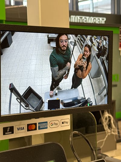 The image displays a photograph of a security monitor showing two individuals within a retail store environment, specifically a self-checkout area in Imatrankoski, Imatra, Finland, as indicated by the mirrored text "IMATRANKOSKI" in the background.
On the monitor, a Caucasian man with long dark hair, a beard, and eyeglasses is positioned on the left. He is wearing a dark green t-shirt, revealing heavily tattooed arms, and is flexing his right bicep while looking directly into the camera. To his right, a Caucasian woman with long dark hair is smiling and holding a smartphone, seemingly capturing an image or video. She is dressed in a dark sports bra and dark leggings, exposing her midriff.
The monitor's display shows a gray tiled floor, a self-checkout station with a black shopping basket on a stand, and a small counter containing a pink/red cup and a white bottle. Shelves stocked with products are visible in the background, further indicating a retail setting.
Below the monitor, a white strip details accepted payment methods, including "BUSINESS" (with an 'S' logo), "VISA", "Mastercard", "AMERICAN EXPRESS", "G Pay", and "Apple Pay". Black cables are visible in the immediate foreground, part of the equipment setup.
