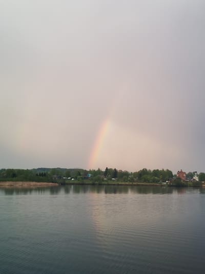 The image captures a tranquil scene in Анхимово, Russia, showing a body of water, presumably a lake or river, under an overcast sky. A faint rainbow arcs across the sky, its ends appearing to touch the horizon.
The foreground is dominated by the water, displaying gentle ripples. The middle ground features a strip of land with lush green vegetation, including trees and reeds. Several buildings are visible nestled among the trees, including what appears to be a brick building and a church with a white spire.
The background is a muted sky filled with a soft, diffused light, contributing to a peaceful atmosphere. No people are visible in the scene. The interaction between the elements – water, land, sky, and rainbow – creates a serene natural landscape.