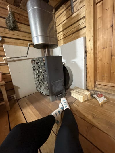A first-person, overhead shot shows a sauna interior with wood-paneled walls and flooring. The main feature is a metal sauna stove, filled with stones. A cylindrical water tank is connected to the stove. A bundle of sauna whisks lies on the floor. A pair of legs clad in black leggings and patterned socks are present. Wood blocks and a sauna glove are on the floor. The scene appears to be inside a private sauna. The location is presumed to be Bashur, Russia.