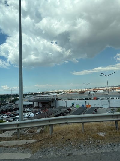 A medium-shot image captures a wide view of a modern commercial district under a partly cloudy sky in Efkarpia Municipal Unit, Greece. A tall, grey pole stands prominently in the left foreground. Beyond it, a large building complex with a metallic facade and large windows stretches across the middle ground. The building features prominent signage, including "SKLAVENITIS" in large white letters. Rows of cars are parked in a vast lot in front of the building, with some covered parking structures visible. The scene is framed by a metal guardrail in the lower foreground, with dry grass and gravel. In the distance, a dense urban landscape with buildings and rooftops extends to the horizon, partially obscured by a haze. Several streetlights are visible throughout the scene. The sky is a blend of blue with scattered white and grey clouds.