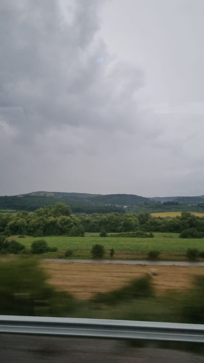 The image depicts a landscape view from a moving vehicle, likely a train or car, in or near the city of Studena, Bulgaria. The scene is dominated by a cloudy, overcast sky, with faint, blurry blue lights visible in the upper portion of the frame, possibly reflections or atmospheric phenomena. Below the sky, a rolling landscape unfolds, featuring a line of trees that transition into further hills and mountains in the distance. In the middle ground, there are fields of green grass and yellow flowers, interspersed with patches of trees. A road or path runs horizontally across the mid-ground. The foreground shows a blurred field of dry grass or hay, possibly with hay bales, and a metallic guardrail runs along the bottom edge of the image, indicating the vantage point is from a road or elevated position. The overall impression is a rural, natural setting under a subdued, grey sky.