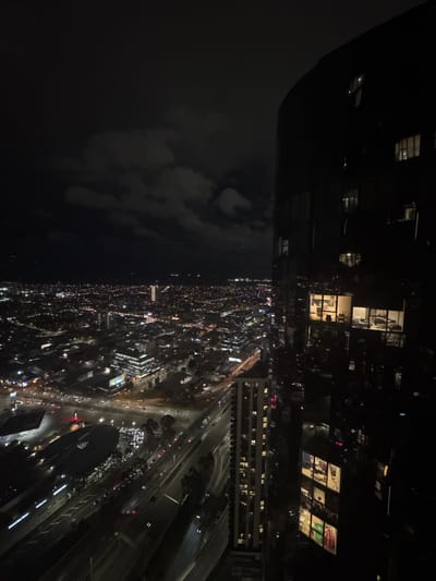 This is a nighttime aerial photograph of the city of Southbank, Australia. On the right side of the frame, a dark, curved skyscraper dominates the foreground, its glass facade reflecting the city lights and revealing illuminated interior spaces with furniture and what appear to be office desks. To the left of the skyscraper, a sprawling urban landscape unfolds, a tapestry of countless lights from buildings, streets, and vehicles stretching into the distance. Several other buildings of varying heights are visible throughout the cityscape, some with illuminated windows. A multi-lane highway, with visible streaks of light from moving cars, cuts through the lower left portion of the image, bordered by trees and more buildings. The sky above is dark with scattered clouds, some of which are faintly illuminated by the city glow. The overall impression is one of a vibrant, active metropolis at night.