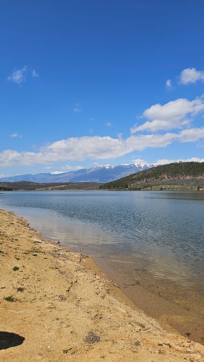 The image depicts a landscape with a lake, mountains, sky, and shoreline. On the left side of the image, the sky is blue with scattered white clouds. In the middle, a mountain range is visible, with some snow on the peaks. A line of trees runs along the base of the mountains, near the lake. The lake is present in the middle of the image, with a visible shoreline on the right. The shoreline is sandy and slopes downward toward the water.
Given the location of Domlyan, Bulgaria, and the terrain depicted, this could be a lake and mountain landscape in the region.