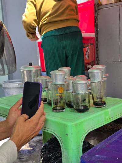 In Binh Thuan Ward, Vietnam, a person is holding a purple smartphone in their hands, partially obscuring a row of Vietnamese coffee filter setups. These setups consist of small metal filter devices stacked on top of clear glass cups, each containing a dark liquid, presumably coffee. The cups are arranged on a bright green plastic table. Behind the table, a person wearing a mustard-yellow sweatshirt and dark green pleated pants is visible, seemingly engaged in preparing or serving something. To the right, a large red container and a metal chain are noticeable. The ground is concrete, with a black trash bag visible beneath the table.