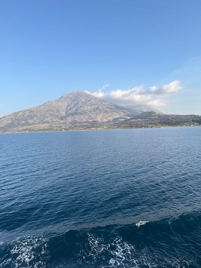 The image displays a panoramic marine landscape near Kamariotissa, Greece. The foreground consists of deep blue water with extensive ripples across its surface. A distinct white wake, indicative of a moving vessel, is visible in the lower right quarter of the frame. The midground shows the expanse of the sea extending towards a distant landmass. The background is dominated by a large, rugged mountain, consistent with Mount Saos, featuring grey, rocky terrain and sparse green vegetation. A narrow strip of greener land with scattered small buildings and structures extends along the mountain's base at the coastline. White cumulus clouds are present, partially obscuring the higher peaks on the right side of the mountain. A tall mast or tower is discernible on a lower hill to the far right. The sky above is a clear, bright blue. No individuals or active interactions between entities are visible.