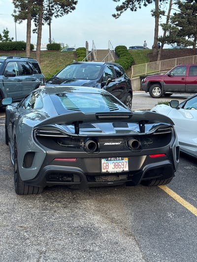 The image shows the rear of a gray McLaren sports car parked in a parking lot. The license plate reads "FIELDS GB 38697" and "LAND OF LINCOLN, Orland Park" (Illinois). Several other vehicles, including a red truck and a white car, are visible nearby. The location appears to be an outdoor parking lot with trees and landscaping in the background. The image does not provide any identifiable features or locations in Oak Brook, IL.