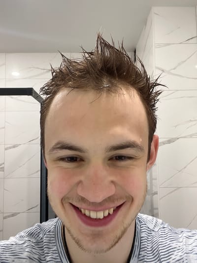 The image displays a young adult male in what appears to be a bathroom setting within Tliustenkhabl', Russia. The subject has wet, spiky, dark brown hair with visible water droplets on his forehead and hair strands. His complexion is fair, and he possesses dark eyes. He is smiling broadly, revealing his upper and lower teeth, and is wearing a light-colored t-shirt with thin, dark horizontal stripes. The background consists of white rectangular wall tiles featuring grey veining, consistent with a marble pattern. A dark, vertical structural element, potentially part of a shower enclosure, is partially visible on the left side of the frame. The subject is looking directly towards the camera.