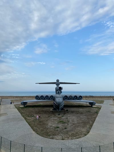 In Arablyar, Russia, a large, gray, Soviet-era ekranoplan (ground-effect vehicle), the Lun-class Ekranoplan, is displayed on a grassy embankment overlooking the Caspian Sea. The vehicle is oriented front-facing, with its twelve engines mounted in two rows of six on the wings. Its distinctive fuselage, designed for low-altitude flight over water, is prominent. A pathway of light-colored interlocking pavers leads from the foreground, bordered by a dark metal fence, towards the ekranoplan. In the middle ground, several small figures, possibly tourists, are visible near the ekranoplan. Beyond the vehicle, a sandy beach stretches along the shoreline of the calm, blue sea. The sky above is a soft blue, with scattered white clouds.