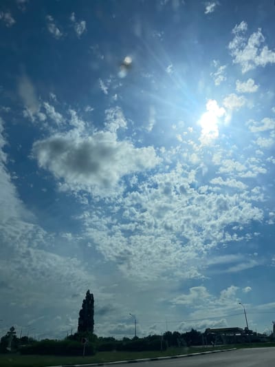 The image shows a bright, sunny sky with scattered clouds. The sun is the dominant subject, shining brightly in the upper left corner with visible rays and lens flare. The clouds range from wispy cirrus formations to larger cumulus clouds. In the foreground, the scene appears to be taken from a moving vehicle, indicated by the blurred elements and the perspective.
To the right, a road or paved surface is visible, bordered by a grassy area and a line of trees. There are several streetlights and a traffic sign with a red arrow on it, suggesting an urban or suburban environment. A tall, dark, conical tree stands out against the sky and clouds. The overall impression is of a daytime scene, possibly late morning or early afternoon, with fair weather. The presence of a road, streetlights, and traffic signs points to a location in a developed area. The image is oriented vertically, as if the camera was held upright. There is no visible text in the image.